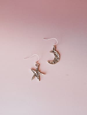 A pair of mismatched silver drop earrings — one star, one crescent moon — laid against a soft dusty rose background, with that worn, slightly antiqued finish that feels like it came from somewhere meaningful.