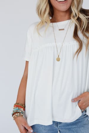 A woman in the Sunday Morning Babydoll Top - White, styled with blue jeans, layered necklaces, and colorful beaded bracelets on her left wrist, stands smiling with one hand in her pocket.