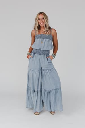 A woman with long blonde hair smiles against a plain background, wearing the Harlow Wide Leg Chambray Jumpsuit in blue. The strapless boho jumpsuit features tiered wide legs and is styled with colorful bracelets and a necklace.