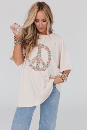 A blonde woman wears the Jude Oversized Peace Sign Graphic Tee in oatmeal, with a colorful floral graphic, paired with light blue jeans and beaded bracelets. She looks down, holding her hair for a relaxed boho vibe.
