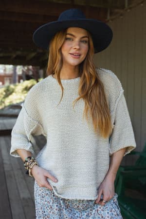 A woman with long, reddish hair wears the Crisp Air Sweater - Oatmeal, a wide-brimmed black hat, a patterned skirt, and bracelets while standing on a wooden porch with greenery in the background.
