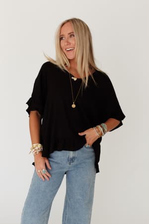A woman with long blonde hair smiles while looking away, wearing The Nest Let Me Be Ruffled Top in black and light blue jeans, accessorized with layered necklaces, bracelets, and rings against a plain white background.