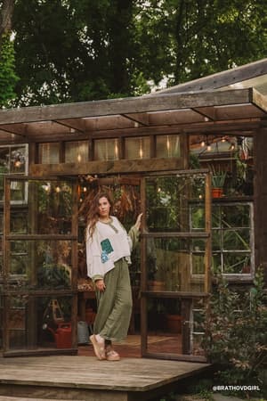 A woman with long hair stands before a rustic greenhouse surrounded by greenery. She wears a loose white top and the So Comfy Wide Leg Pant Cropped Length in Olive, with sandals, posing with one hand on the doorframe.