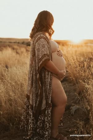 A pregnant woman stands in a sunlit field at sunset, gently holding her belly while wearing the Paisley Tapestry Free Flow Duster Kimono - Brown. Tall golden grass surrounds her, highlighting the soft elegance of her kimono.