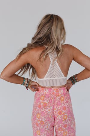 A woman with long, wavy blonde hair stands with her back to the camera, wearing the Infinite Expressions Bralette - Ivory and high-waisted pink floral pants. Colorful beaded bracelets adorn both of her wrists.