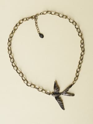 A bronze-toned chain necklace with a detailed flying bird pendant — the kind of piece that feels like it has a story, laid flat against a soft cream background.