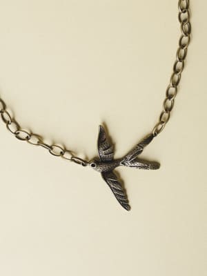 A close-up of a bronze bird-in-flight pendant necklace laid on a warm cream surface — the kind of piece that feels like it has a little story behind it.