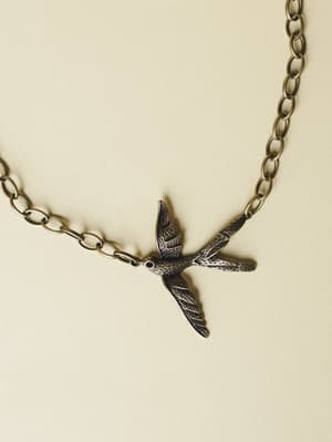 A close-up of a bronze bird-in-flight pendant necklace laid on a warm cream surface — the kind of piece that feels like it has a little story behind it.