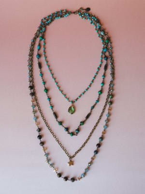 Four beaded necklaces layered together on a blush pink surface — turquoise and mixed stone beads on antique brass chains, anchored by a green teardrop stone pendant and a small gold star charm.
