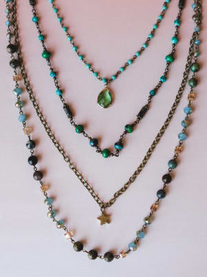 Three layered beaded necklaces arranged on a blush background — one with tiny turquoise stones and a green teardrop pendant, one with mixed earth-tone beads, and a longer brass chain with a small star charm.