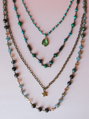 Three layered beaded necklaces arranged on a blush background — one with tiny turquoise stones and a green teardrop pendant, one with mixed earth-tone beads, and a longer brass chain with a small star charm.