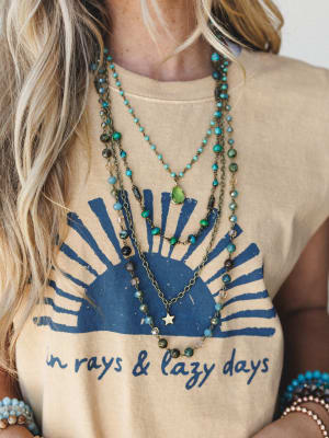 A close-up torso shot layering boho beaded necklaces — turquoise, green stone, and gold star pendants — styled over a sandy tan graphic tee that reads 'in rays & lazy days,' with stacked bead bracelets peeking in at the edges.