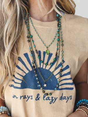 A close-up torso shot layering boho beaded necklaces — turquoise, green stone, and gold star pendants — styled over a sandy tan graphic tee that reads 'in rays & lazy days,' with stacked bead bracelets peeking in at the edges.