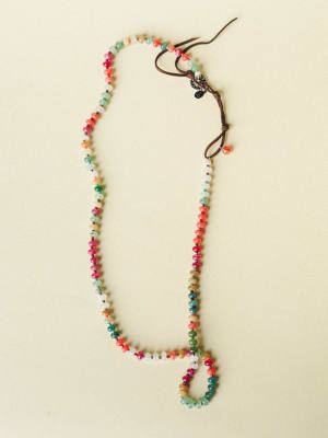 A long, colorful beaded necklace with multicolor gemstone-style rondelle beads laid flat against a warm cream background — the kind of piece that looks like it collected a little bit of everywhere.