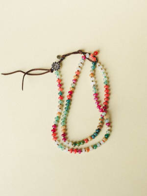 A long multicolor beaded necklace coiled loosely on a soft cream background, featuring rondelle stone beads in a confetti mix of colors with a leather tie closure and a small silver daisy charm.