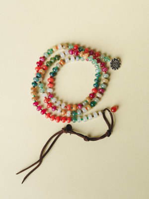 A colorful multi-strand beaded wrap bracelet or long necklace coiled on a soft cream surface, featuring mixed stone beads in warm and cool tones with a silver flower charm and brown suede tassel closure.