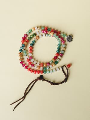 A colorful multi-strand beaded wrap bracelet or long necklace coiled on a soft cream surface, featuring mixed stone beads in warm and cool tones with a silver flower charm and brown suede tassel closure.