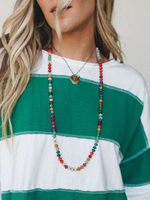 A close-up lifestyle shot of a long multicolor beaded stone necklace layered with a gold coin charm necklace, styled over a green and white colorblock long-sleeve top with contrast stitching.