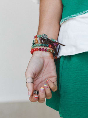 A close-up of a stacked beaded bracelet set worn on the wrist, layered with a leather wrap and silver flower charm — colorful, personal, and very much the kind of jewelry that tells a story without saying a word.