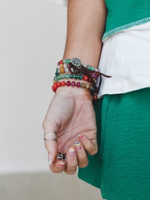 A close-up of a stacked beaded bracelet set worn on the wrist, layered with a leather wrap and silver flower charm — colorful, personal, and very much the kind of jewelry that tells a story without saying a word.