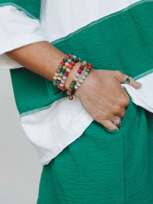 A close-up of a stacked beaded bracelet set worn at the wrist, layered in earthy multicolor stones against the contrast of a white-and-green color-blocked top — the kind of detail shot that makes you want to reach for every bracelet you own.