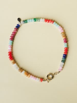 A multicolor beaded necklace laid flat on a soft cream surface — rondelle-cut stones in every color of the rainbow with a gold spring-ring clasp, the kind of piece that looks like it's been collected bead by bead over a lifetime of happy days.