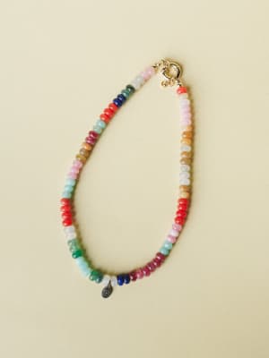 A multicolor gemstone beaded necklace laid flat against a soft sage background, with rondelle-cut beads in every color of the rainbow and a gold spring ring clasp — the kind of piece that makes a plain white tee feel like a whole personality.