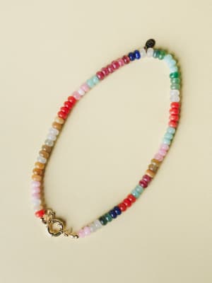 A multicolor beaded necklace laid flat against a warm cream background — rondelle-shaped stones in a full rainbow of hues with a polished gold spring ring clasp, the kind of piece that looks like it has a story.