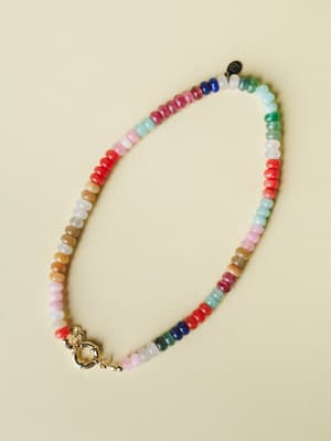 A multicolor beaded necklace laid flat against a warm cream background — rondelle-shaped stones in a full rainbow of hues with a polished gold spring ring clasp, the kind of piece that looks like it has a story.