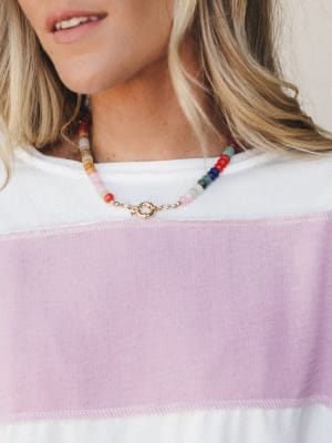 A close-up of a multicolor beaded necklace with a gold spring-ring clasp, worn over a white and lavender color-block top — playful and personal, like it came straight out of a jewelry box you've been collecting since forever.