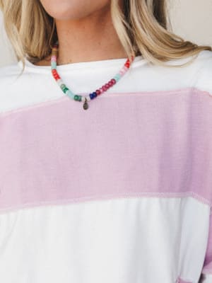 A close-up of a colorful beaded necklace with a small charm pendant, layered over a wide-stripe white and lilac top — playful and personal, like something you'd never want to take off.