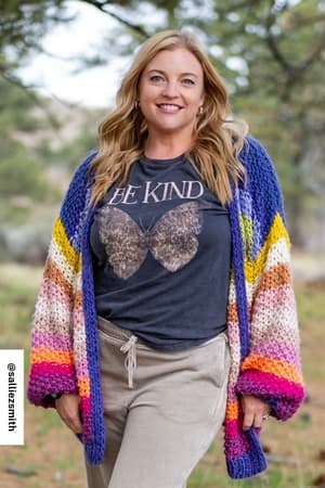 A smiling woman stands outside wearing the Be Kind Mineral Washed Graphic Tee - Charcoal with a butterfly and “Be Kind” motif, paired with a colorful knit cardigan and beige pants. Greenery and trees blur in the background.