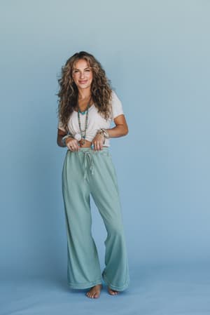 A woman with long curly hair smiles barefoot against a light blue background, wearing layered necklaces, bracelets, a white cropped t-shirt, and the So Comfy Wide Leg Pant Cropped Length in sage with a drawstring waist.