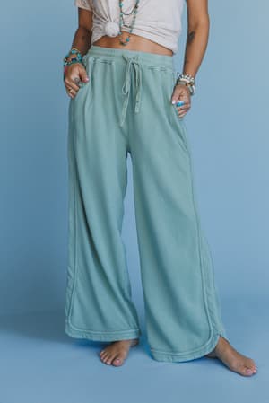 A person stands barefoot on a blue floor, wearing So Comfy Wide Leg Pant Cropped Length in Sage with a drawstring waist and a knotted white t-shirt. Colorful bracelets and tattoos highlight their arms.