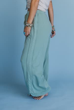 A person stands barefoot against a blue background, showing only the lower half of their body dressed in So Comfy Wide Leg Pant Cropped Length - Sage, paired with a white top and accessorized with layered bracelets and rings.
