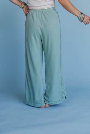 A person stands barefoot against a matching blue background, wearing So Comfy Wide Leg Pant Cropped Length in Sage with a drawstring waist, several bracelets, one hand on their hip, and the other slightly raised.
