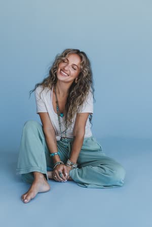 A woman with long wavy hair sits barefoot against a blue backdrop, smiling with eyes closed. She wears a white t-shirt, layered jewelry, and the So Comfy Wide Leg Pant Cropped Length in sage with a drawstring waist.