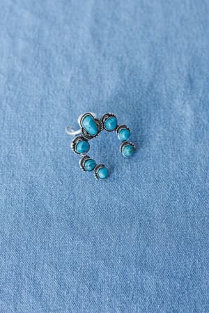 The Celia Blossom Turquoise Ring features seven oval turquoise stones in a crescent design on a silver tone band, showcased on light blue denim fabric.