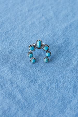 The Celia Blossom Turquoise Ring features seven oval turquoise stones in a silver-tone, fan-shaped design, displayed on light blue denim fabric.