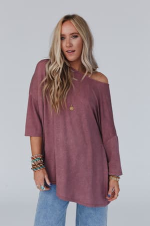 A woman with long blonde hair wears The Nest Sunday Stroll Oversized Tee in mauve styled off-the-shoulder, light blue jeans, layered necklaces, and several bracelets, standing against a plain light background.