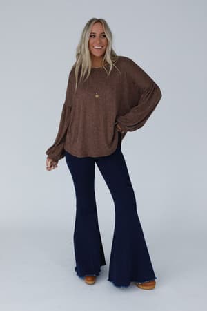 A woman with long blonde hair smiles, wearing The Nest Sweet Heart Balloon Sleeve Knit Sweater in Mocha, dark blue flared jeans, brown shoes, and a gold pendant necklace against a plain light background.