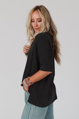 A woman with long blonde hair wears The Nest On The Go V Neck Ribbed Essential Tee in Charcoal, styled with light blue jeans and colorful beaded bracelets as she poses in profile against a plain white background.