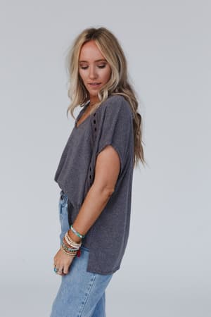 A woman with long, wavy blonde hair stands sideways and looks down, wearing The Nest Camden Lace V Neck Top in charcoal and light blue jeans. Colorful bracelets accent her left wrist against a plain light background.