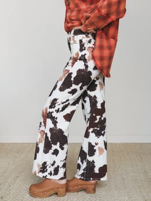 A side-profile shot of wide-leg cow print pants paired with a knotted fringe-hem plaid top and studded wood clogs — the kind of outfit that means business at a country concert but still feels totally like *you*.