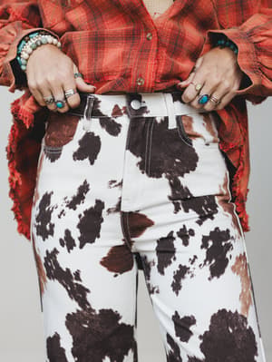 A close-up torso shot of cow-print jeans paired with a frayed plaid flannel shirt, styled with stacked silver rings, turquoise stones, and beaded bracelets — western-boho in the best possible way.