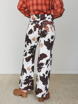 Shot from behind, a model wears wide-leg cow print jeans paired with a fringed plaid top and studded clogs — a full western moment that somehow feels totally wearable.
