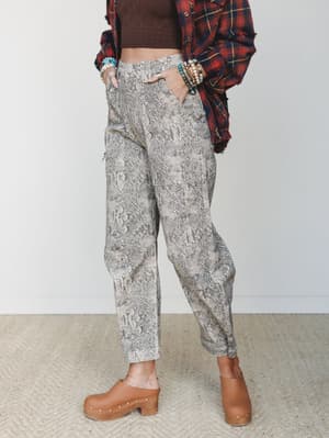 A mid-shot styled look centered on barrel-leg snakeskin print jeans, layered with a dark crop top and open plaid flannel shirt, finished with tan studded clogs and stacked beaded bracelets — lived-in and expressive with an effortless earthy edge.