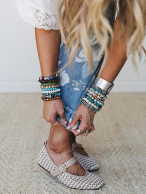 A close-up lifestyle shot of a model bending to adjust her woven flats, wrists stacked with beaded and cuff bracelets, the focus pulling between the floral denim at her knees and the layered jewelry — the kind of detail shot that makes you want to st