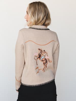 A blonde model shot from behind shows off the standout detail of this oatmeal knit sweater — a whimsical embroidered cowgirl riding a rearing horse across the back, finished with contrast black blanket-stitch trim at the hem and collar.