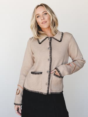 A knit cardigan jacket in warm oatmeal with bold black whipstitch trim, silver concho buttons, and embroidered cuff details — the kind of piece that feels like a heirloom you'd actually wear on a Tuesday.